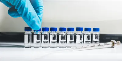 A gloved hand arranges small labeled vials containing liquid samples on a laboratory bench next to a syringe.