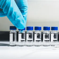 A gloved hand arranges small labeled vials containing liquid samples on a laboratory bench next to a syringe.
