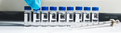 A gloved hand arranges small labeled vials containing liquid samples on a laboratory bench next to a syringe.