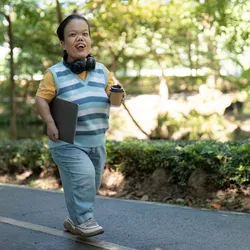 Young individual with dwarfism smiling while walking in a sunny park holding a tablet.