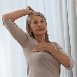 Senior woman doing breast self-examination near mirror indoors.