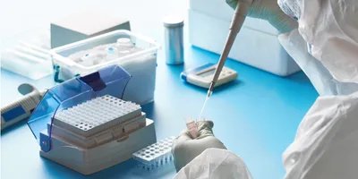 Scientist in protective laboratory clothing using a pipette to transfer liquid into a sample tube beside a PCR tube rack and laboratory equipment.