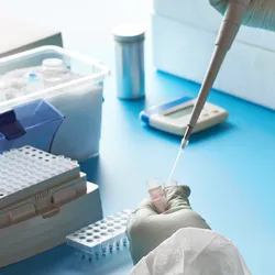 Scientist in protective laboratory clothing using a pipette to transfer liquid into a sample tube beside a PCR tube rack and laboratory equipment.