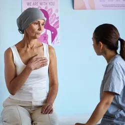 A patient with breast cancer sitting on a medical bed while talking to a nurse.