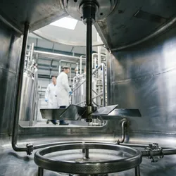 A view from inside a large, polished stainless steel industrial mixing vessel, showing a central agitator and bottom sparger, with a facility and two workers visible in the distance.