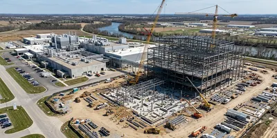 Aerial photograph of a pharmaceutical GMP manufacturing facility under construction, with steel framing of the new building visible beside a completed existing facility.