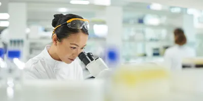 A scientist in a white lab coat looking into a microscope in a brightly lit modern laboratory.