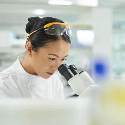 A scientist in a white lab coat looking into a microscope in a brightly lit modern laboratory.