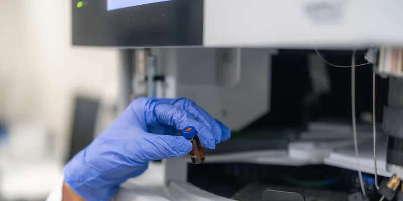 A researcher wearing a white lab coat and blue nitrile glove holds a tiny brown glass sample vial with a blue cap in front of an autosampler carousel on a HPLC laboratory analyzer.