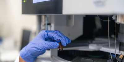 A researcher wearing a white lab coat and blue nitrile glove holds a tiny brown glass sample vial with a blue cap in front of an autosampler carousel on a HPLC laboratory analyzer.