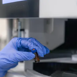 A researcher wearing a white lab coat and blue nitrile glove holds a tiny brown glass sample vial with a blue cap in front of an autosampler carousel on a HPLC laboratory analyzer.