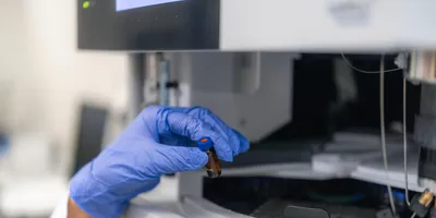 A researcher wearing a white lab coat and blue nitrile glove holds a tiny brown glass sample vial with a blue cap in front of an autosampler carousel on a HPLC laboratory analyzer.