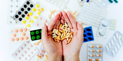 Different medications and pill packets on a table and a pair of hands cupping a handful of pills. 