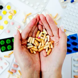 Different medications and pill packets on a table and a pair of hands cupping a handful of pills. 