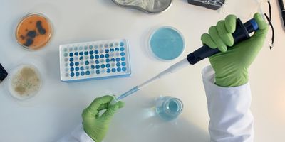 Top view of a laboratory researcher wearing green gloves and using a pipette to transfer liquid at a bench surrounded by other laboratory equipment.