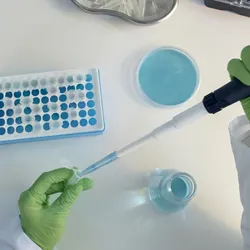 Top view of a laboratory researcher wearing green gloves and using a pipette to transfer liquid at a bench surrounded by other laboratory equipment.