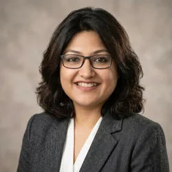 A headshot of Priyom Bose wearing a gray suit jacket on a light brown background