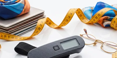 A studio stock photograph on a plain white surface featuring a yellow measuring tape curving and coiling dynamically around various objects. In the center sits a small, black carbon-fiber electronic device with a digital display. Surrounding it are a pair of wire-rimmed reading glasses, stacked notebooks, and folded blue and orange patterned fabric scarves. JPG + 2