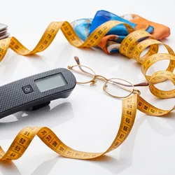 A studio stock photograph on a plain white surface featuring a yellow measuring tape curving and coiling dynamically around various objects. In the center sits a small, black carbon-fiber electronic device with a digital display. Surrounding it are a pair of wire-rimmed reading glasses, stacked notebooks, and folded blue and orange patterned fabric scarves.    JPG + 2
