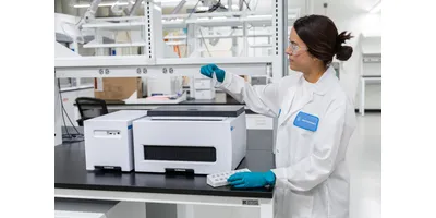  Researcher in white lab coat and teal gloves holding a sample vial next to a benchtop oligonucleotide synthesizer instrument in a modern laboratory.