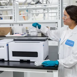  Researcher in white lab coat and teal gloves holding a sample vial next to a benchtop oligonucleotide synthesizer instrument in a modern laboratory.